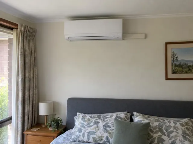 Wall-mounted split system above a bed in a carpeted bedroom with curtains and bedside table in Craigmore